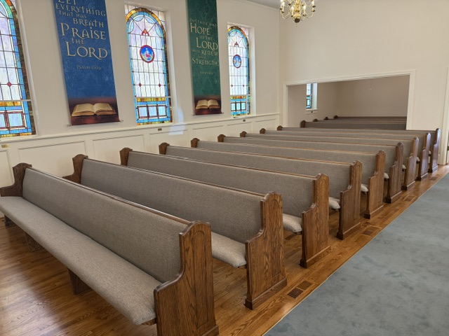 Rows of restored and reupholstered oak church pews on refinished hardwood floors beneath arched stained-glass windows